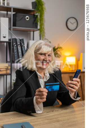 Happy Caucasian senior woman using credit card and smartphone for online shopping payments at office Happy Caucasian senior woman using credit card and smartphone for online shopping payments at office 125676988