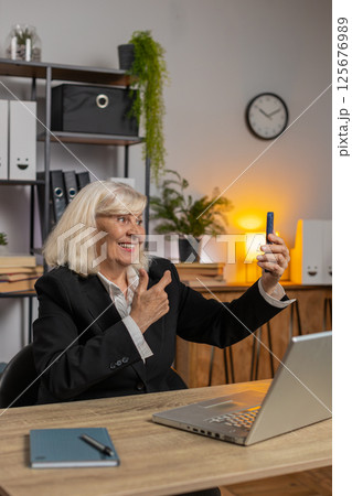 Senior Caucasian businesswoman waves hand greeting welcomes on smartphone at home office desk 125676989