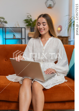Happy young woman in dress sitting on sofa with laptop working on project in living room at home Happy young woman in dress sitting on sofa with laptop working on project in living room at home 125677015