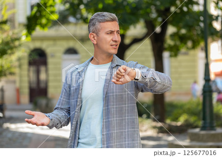 Mature man looking at wristwatch while waiting for someone feeling upset about being late in city 125677016