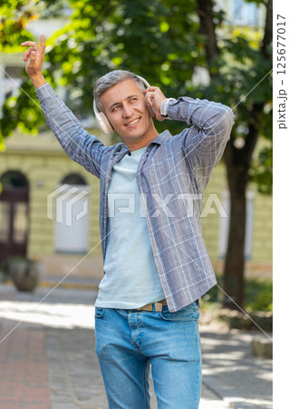 Young gray-haired man listening to music using wireless headphones and smartphone on city street 125677017