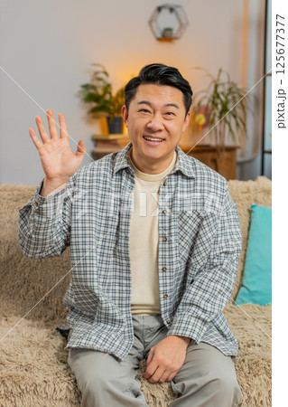Happy young man vlogger smiling friendly at camera and waving hands while sitting on sofa at home 125677377