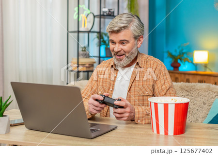 Mature man sitting at table with laptop and gamepad, playing video games and eating popcorn at home 125677402