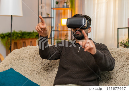 Indian man on sofa wears VR headset at home and reacts with excitement to immersive virtual world 125677467
