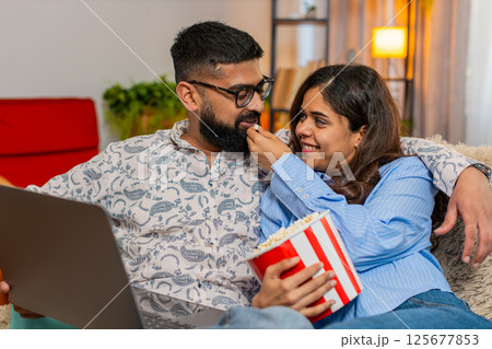 Indian family couple man woman hugging while watching series film on laptop with popcorn, enjoying Indian family couple man woman hugging while watching series film on laptop with popcorn, enjoying 125677853