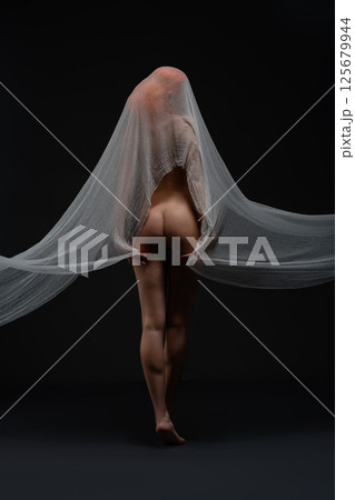 Elegant woman in sheer fabric poses nude against a dark background Elegant woman in sheer fabric poses nude against a dark background 125679944