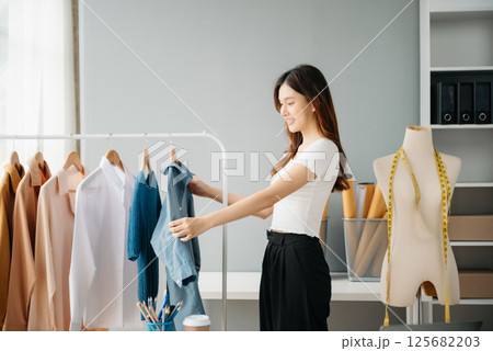Successful fashion designer. Attractive young asian woman with smile while standing in workshop. Beautiful owner business woman working and holds tablet, laptop and smartphone in studio. Successful fashion designer. Attractive young asian woman with smile while standing in workshop. Beautiful owner business woman working and holds tablet, laptop and smartphone in studio. 125682203