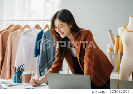 Successful fashion designer. Attractive young asian woman with smile while standing in workshop. Beautiful owner business woman working and holds tablet, laptop and smartphone in studio. 125682205