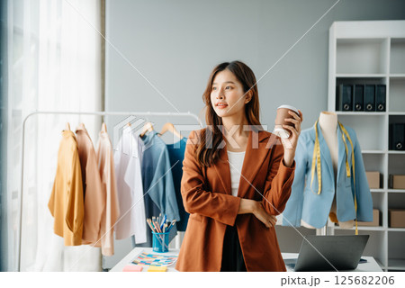 Successful fashion designer. Attractive young asian woman with smile while standing in workshop. Beautiful owner business woman working and holds tablet, laptop and smartphone in studio. Successful fashion designer. Attractive young asian woman with smile while standing in workshop. Beautiful owner business woman working and holds tablet, laptop and smartphone in studio. 125682206