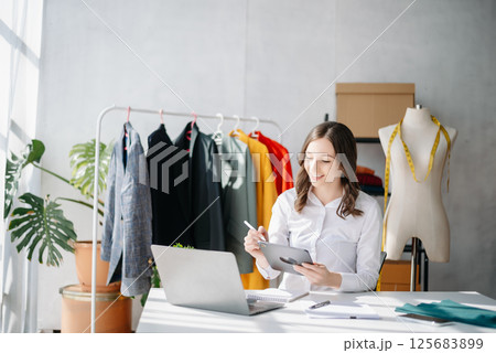 Diverse female fashion designers at work with tailor centimeters on necks and holds tablet and smartphone. Diverse female fashion designers at work with tailor centimeters on necks and holds tablet and smartphone. 125683899