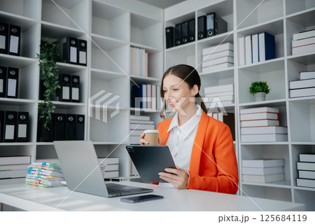 Confident business expert attractive smiling young woman typing laptop ang holding digital tablet on desk in creative office. Confident business expert attractive smiling young woman typing laptop ang holding digital tablet on desk in creative office. 125684119