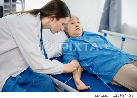 Doctor in white suit take notes while discussing and Asian elderly, woman patient who lying on bed with receiving saline solution in hospital or clinic. 125684453
