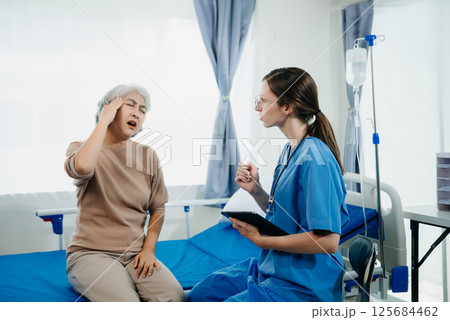 Doctor in white suit take notes while discussing and Asian elderly, woman patient who lying on bed with receiving saline solution in hospital or clinic. 125684462