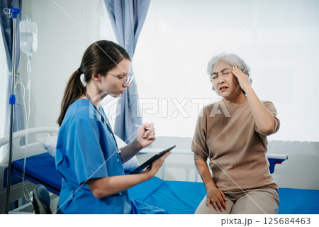 Doctor in white suit take notes while discussing and Asian elderly, woman patient who lying on bed with receiving saline solution in hospital or clinic. 125684463