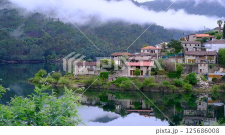 Scenic view of a peaceful mountain village nestled beside a calm lake with perfect reflections of stone houses and forested hills. 125686008
