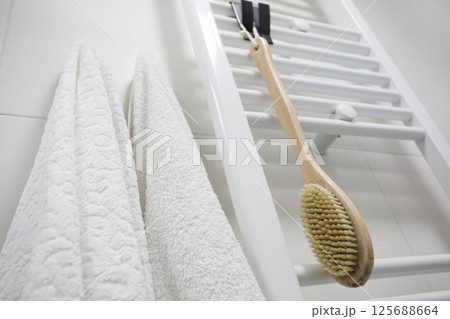 White heated towel rail with white cotton towel and brush for dry brushing massage on wall 125688664