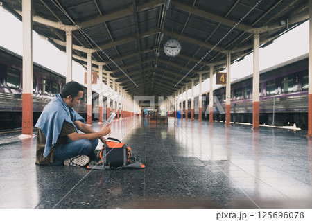 A young man traveler with digital tablet sitting on floor at train station choose where to travel. Concept of backpackers travel adventure by train. 125696078