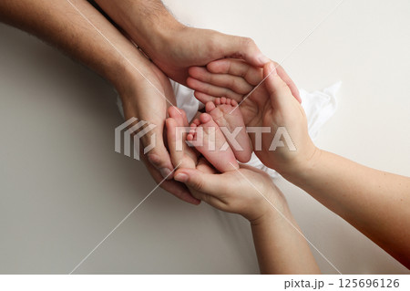 A father and mother hold the feet of a newborn child in a white blanket on a white background.. The feet of a newborn in the hands of parents. Photo of foot, heels and toes.The palms of the parents. 125696126
