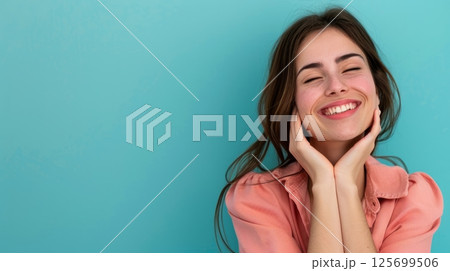 Happy young woman with freckles smiles broadly, hands gently touching her face, against a teal background 125699506
