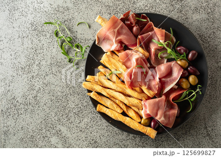 Prosciutto with bread sticks, olives, and rosemary on a black plate. Prosciutto with bread sticks, olives, and rosemary on a black plate. 125699827