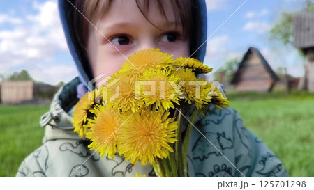 Cute Little Boy Picks Flowers In A Field On A Background Of Sky. Child looks at the dandelion flowers and smiles. small child, boy with yellow flowers 125701298
