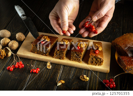 A baker arranges four slices of moist cake on a wooden platter, adding red berries and walnuts as a garnish. The rustic kitchen features dark wood and natural lighting A baker arranges four slices of moist cake on a wooden platter, adding red berries and walnuts as a garnish. The rustic kitchen features dark wood and natural lighting 125701355