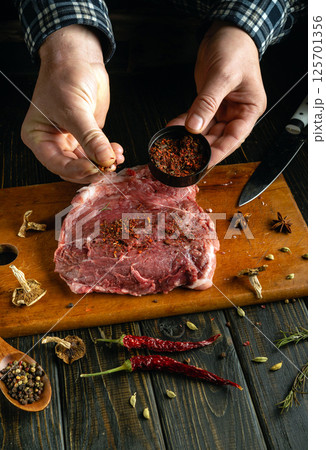 A person seasoned a raw steak with a blend of spices, including dried herbs and chili peppers, on a wooden cutting board in a rustic kitchen. It captures a moment of culinary creativity A person seasoned a raw steak with a blend of spices, including dried herbs and chili peppers, on a wooden cutting board in a rustic kitchen. It captures a moment of culinary creativity 125701356