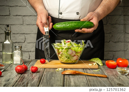 A chef is chopping cucumbers while preparing a fresh salad in a rustic kitchen. Various vegetables and ingredients are spread on the wooden table, creating a vibrant atmosphere A chef is chopping cucumbers while preparing a fresh salad in a rustic kitchen. Various vegetables and ingredients are spread on the wooden table, creating a vibrant atmosphere 125701362