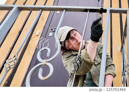 Middle aged woman in light cap and work glove paints vertical metal bars of porch railing with black paint using paintbrush near entrance door. Middle aged woman in light cap and work glove paints vertical metal bars of porch railing with black paint using paintbrush near entrance door. 125701464