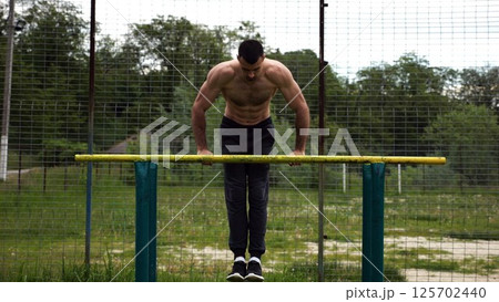 Hardy shirtless sportsman doing exercises on parallel bars at nature. Strong and muscular man working out at sports ground. Young athletic guy exercising outdoor. Concept of active healthy lifestyle Hardy shirtless sportsman doing exercises on parallel bars at nature. Strong and muscular man working out at sports ground. Young athletic guy exercising outdoor. Concept of active healthy lifestyle 125702440