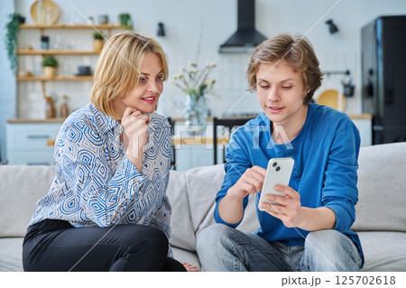 Middle aged mother and teenage son sitting together at home looking at smartphone 125702618