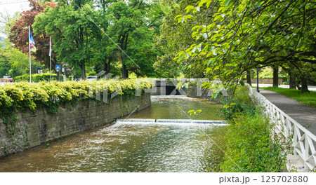 Old waterfall in the summer city with old bridge. Nature background. Falls with clean water in sunny day and green grass around. Old waterfall in the summer city with old bridge. Nature background. Falls with clean water in sunny day and green grass around. 125702880