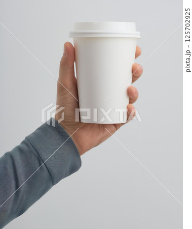 A hand holding a plain white takeaway coffee cup with a lid. The background is minimal and light, highlighting the simplicity and everyday nature of the scene. 125702925