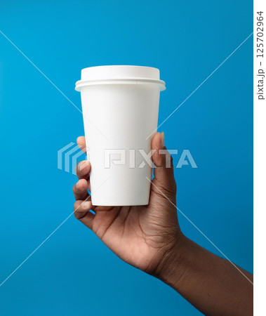 A hand is holding a plain white disposable coffee cup with a lid, set against a bright blue background. The image is simple and clean, focusing on the cup, ideal for branding or advertising 125702964