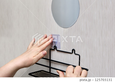 Hands are mounting black wire bathroom shelf on wall using transparent nano adhesive hook for easy and damage-free bathroom hardware fitting 125703131