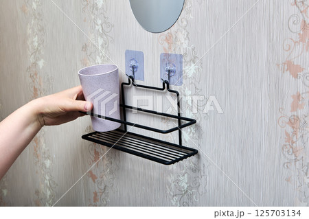 Hand places plastic cup onto black wire bathroom shelf fixed to wall with two transparent nano adhesive hooks for convenient storage solution. Hand places plastic cup onto black wire bathroom shelf fixed to wall with two transparent nano adhesive hooks for convenient storage solution. 125703134