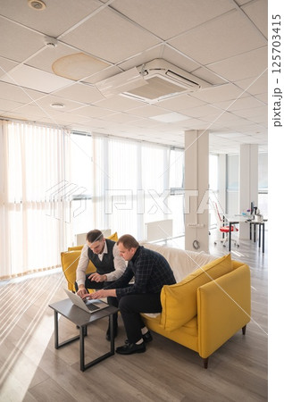 Two Caucasian men discussing work on a laptop while sitting on a sofa. Modern office. Two Caucasian men discussing work on a laptop while sitting on a sofa. Modern office. 125703415
