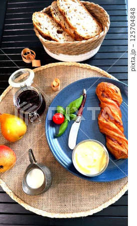 Breakfast spread with golden twisted pastry, basil leaves, cherry tomato, and butter on blue plate, surrounded by basket bread, fresh pears, and jam jar, creating rustic morning mood. 125704848