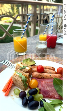Gourmet breakfast on balcony at prestigious tourist destination, sausages and sandwiches artfully arranged, fresh juice highlights leisurely luxury vacation, ideal travel lifestyle capture. Keywords: Gourmet breakfast on balcony at prestigious tourist destination, sausages and sandwiches artfully arranged, fresh juice highlights leisurely luxury vacation, ideal travel lifestyle capture. Keywords: 125704893