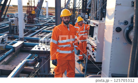 Oil workers in orange uniforms, yellow hardhats checking pump station at sunset, reviewing control panel graphs, examining pipes for operational efficiency 125705312