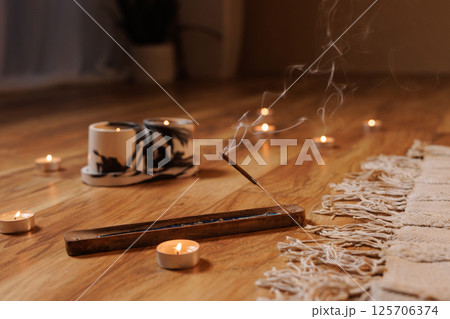 Close-up of burning incense stick with candles on wooden floor, creating a calm, meditative atmosphere for yoga, relaxation, or mindfulness practices. Warm, cozy, and peaceful setting. Close-up of burning incense stick with candles on wooden floor, creating a calm, meditative atmosphere for yoga, relaxation, or mindfulness practices. Warm, cozy, and peaceful setting. 125706374