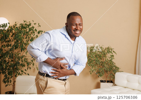 Man suffering from severe stomach pain at home. He clutches his abdomen with a pained expression, indicating digestive issues, cramps, or food poisoning. Health and medical concept. 125706386
