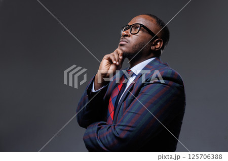 Thoughtful businessman in striped suit and glasses, looking up with hand on chin. Concept of decision-making, planning, strategy, leadership, and innovation. Dark studio background. 125706388