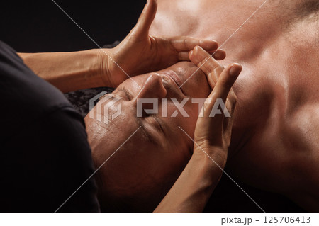 Therapist performing facial massage on a relaxed man in spa setting, focusing on jaw and chin muscle areas. 125706413