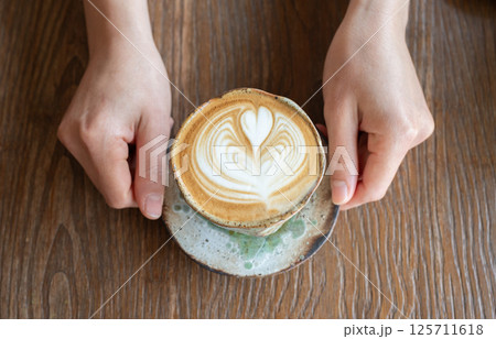 Someone hands holding a cup of hot Cappuccino coffee before drinking. The taste of cappuccinos can be described as creamy, smooth, and balanced. Someone hands holding a cup of hot Cappuccino coffee before drinking. The taste of cappuccinos can be described as creamy, smooth, and balanced. 125711618