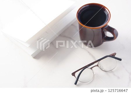 白いテーブルの上の一冊の本とメガネ、コーヒーのマグカップで、コーヒーを飲みながら読書を楽しむイメージ 白いテーブルの上の一冊の本とメガネ、コーヒーのマグカップで、コーヒーを飲みながら読書を楽しむイメージ 125716647