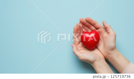 Hands gently holding a small glossy red heart on a light blue background. Symbol of love, health, and care. Hands gently holding a small glossy red heart on a light blue background. Symbol of love, health, and care. 125719736