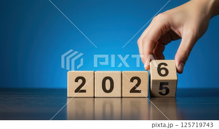 Hand placing a wooden block with number 6 to change year 2025 to 2026 on a blue background. 125719743