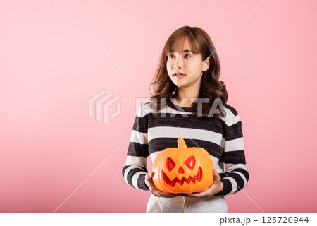 Halloween-themed studio shot, an Asian woman holds a variety of model pumpkins, including a ghost-shaped one, on a pink background. Her cheerful costume sets the tone for a fun and spooky Halloween. Halloween-themed studio shot, an Asian woman holds a variety of model pumpkins, including a ghost-shaped one, on a pink background. Her cheerful costume sets the tone for a fun and spooky Halloween. 125720944
