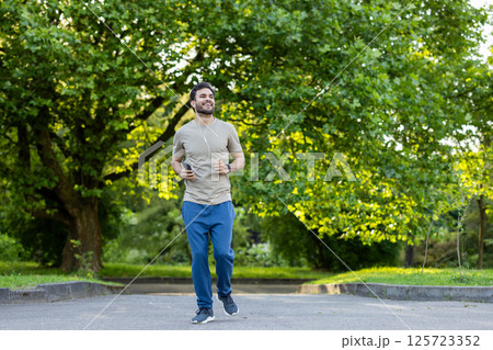 Young man jogging outdoors surrounded by green trees, listening to music. Enjoying fitness and nature during a sunny day. Positive energy and healthy lifestyle concept. 125723352